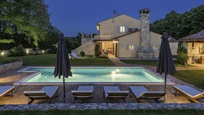 Une piscine avec des chaises longues et des parasols, une maison en pierre dotée d’une cheminée, et un jardin bien éclairé au crépuscule.