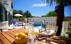A wooden table with a bowl of lemons, a pitcher of water, and a glass of water with a lemon slice. There are lounge chairs and a pool in the background.