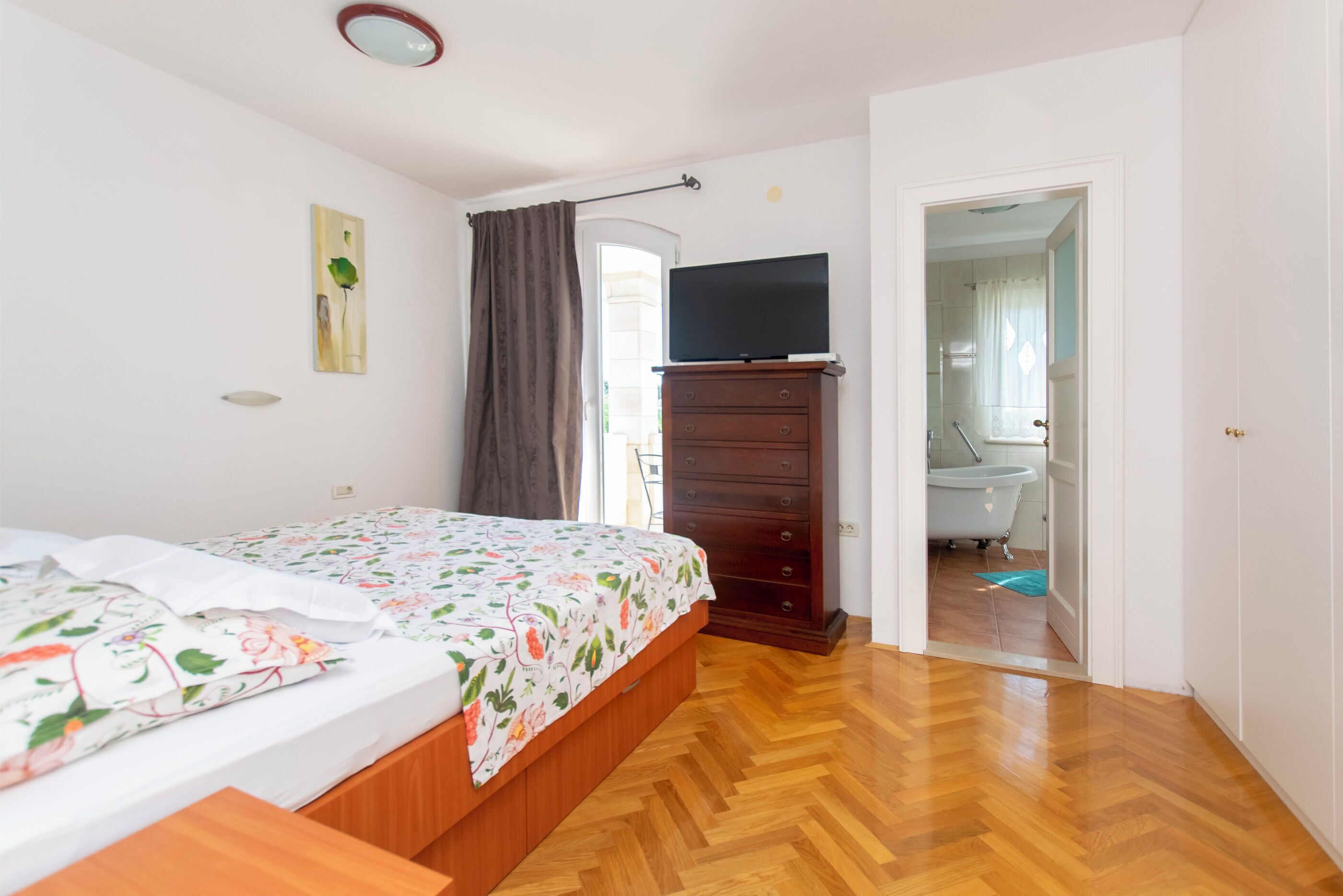 A bedroom with a bed, a dresser, a television, and a bathroom visible through an open door.