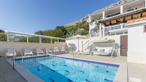 Una piscina con agua azul cristalina, sillas de descanso blancas y una pérgola blanca.