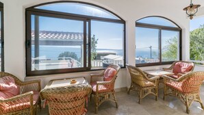 Un balcón soleado con mobiliario de mimbre y amplias ventanas que ofrecen vistas al mar y edificios.