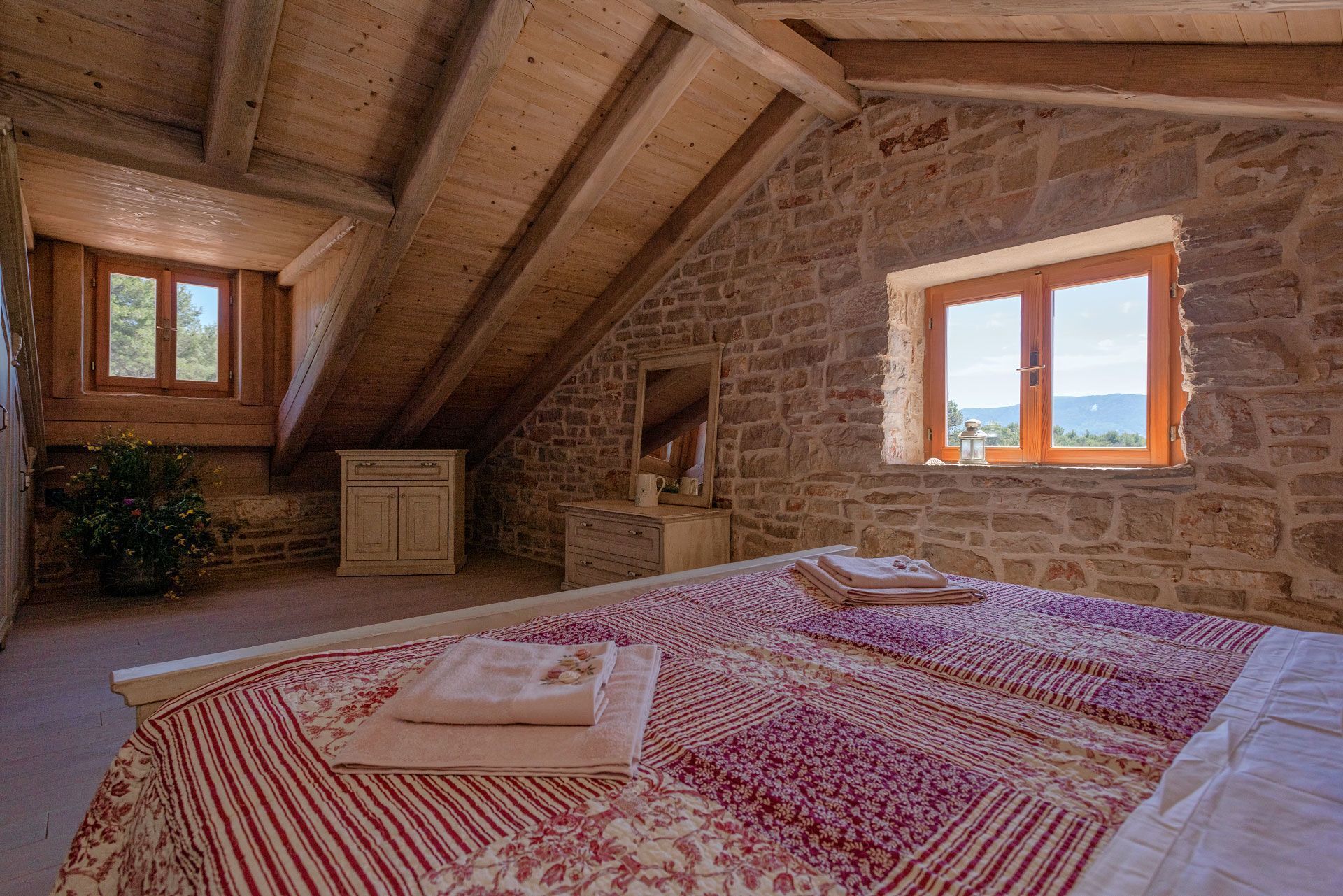 A bedroom with a stone wall, wooden beams, a bed with a floral bedspread, a dresser, and a window with a view.