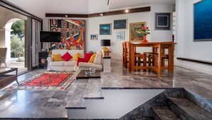 Een ruime woonkamer met een witte bank, kleurrijke kussens, een rood tapijt en een houten eettafel.
