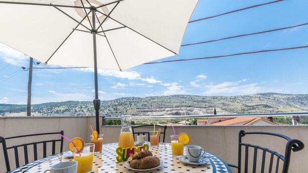 Ein runder Tisch mit weiß-blauen Fliesen, gedeckt mit einem Teller Croissants, Tassen und Gläsern Orangensaft, unter einem weißen Sonnenschirm.