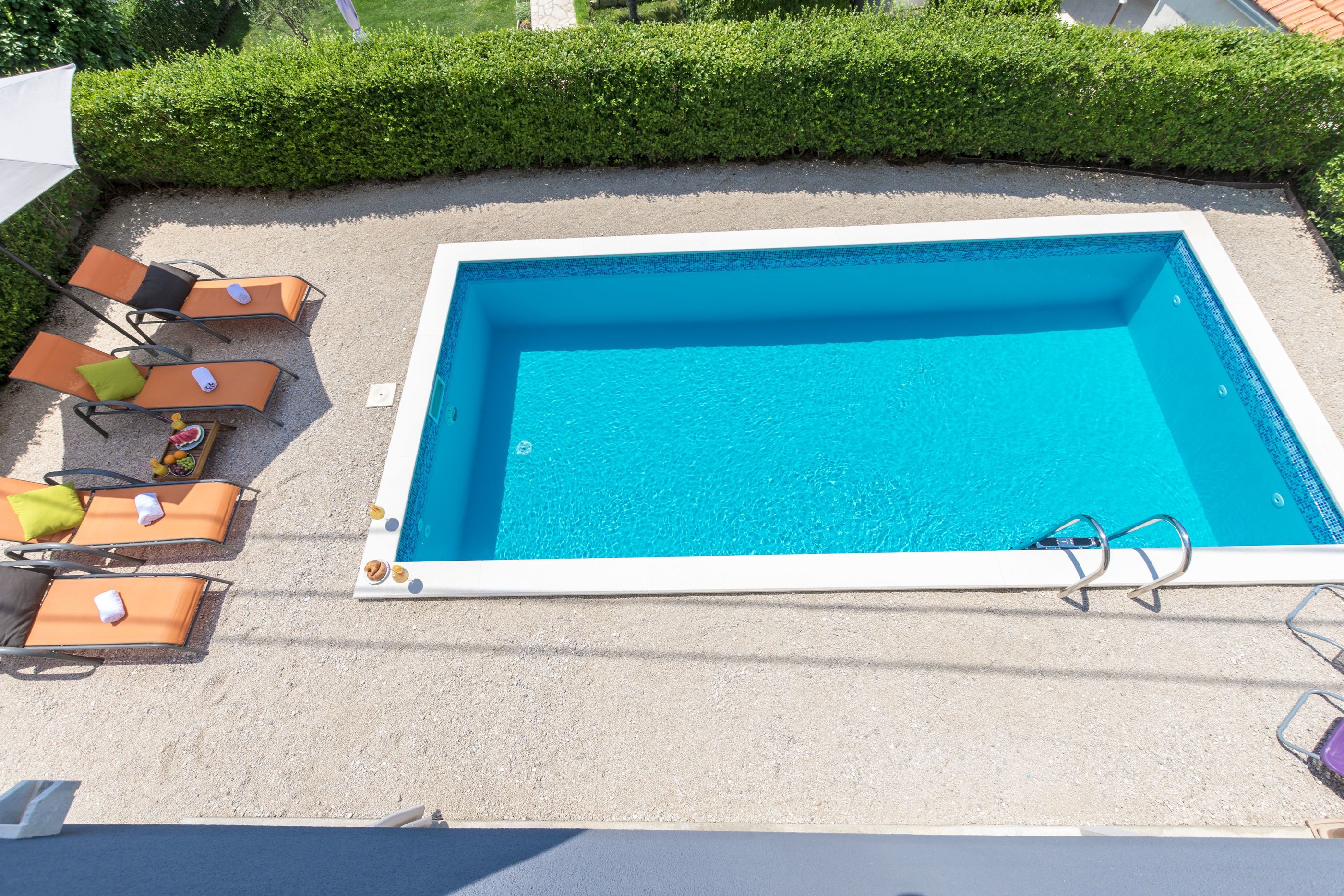 A rectangular swimming pool with a white border, surrounded by lounge chairs and a hedge.