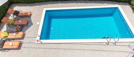 A rectangular swimming pool with a white border, surrounded by lounge chairs and a hedge.