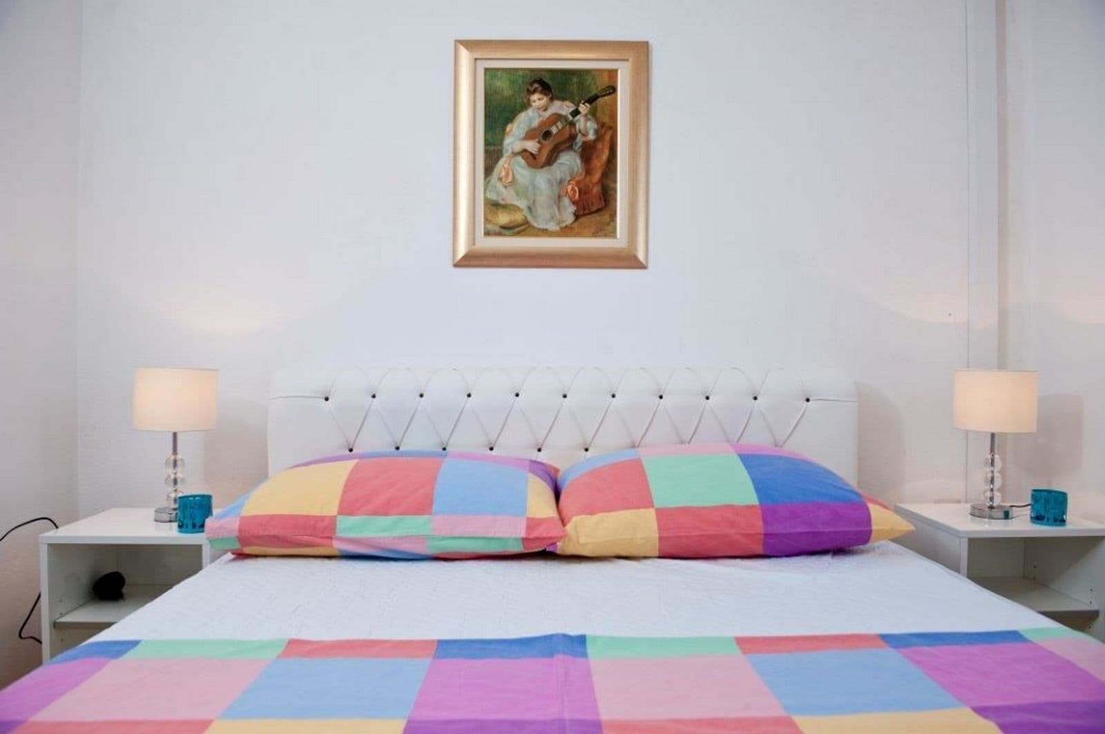 A bedroom with a white bed, colorful checkered pillows, a tufted headboard, and a framed painting above the bed.