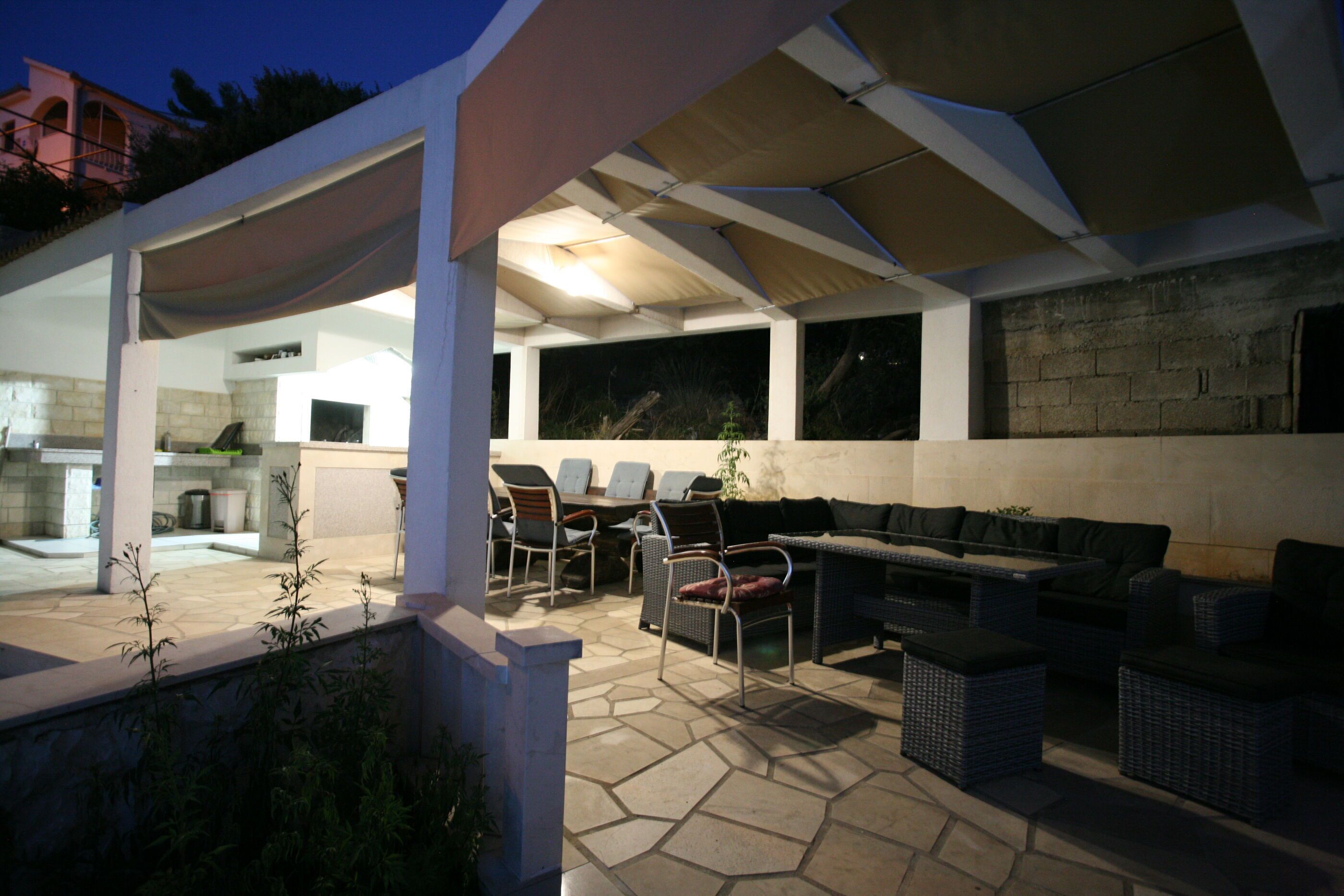 An outdoor patio area with a covered seating area, stone flooring, and a view of the kitchen in the background.