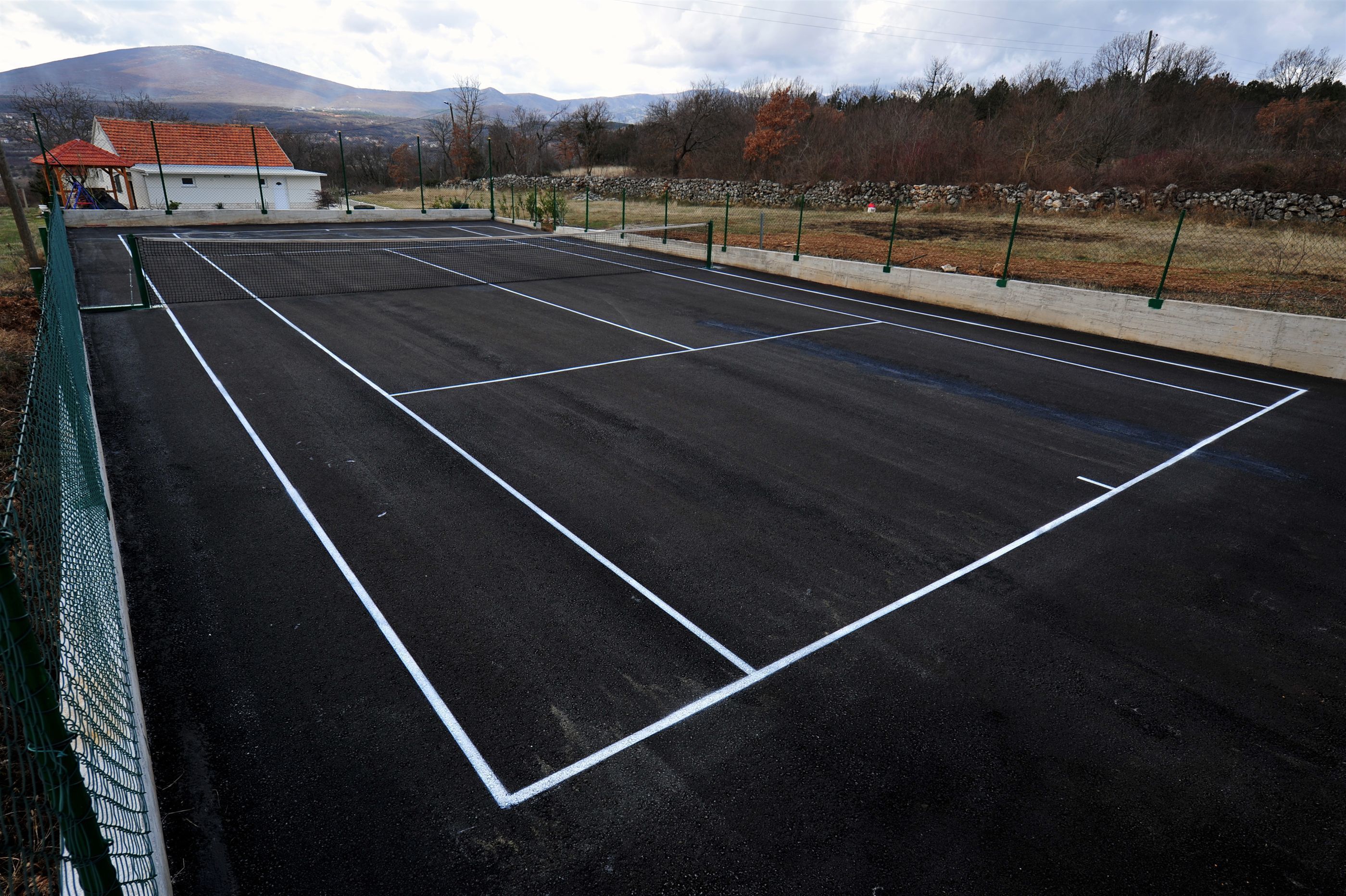 Una pista de tennis amb xarxa, envoltada per una tanca verda, i un edifici petit amb teulada vermella al fons.