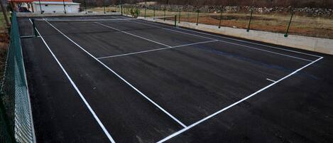 Una pista de tennis amb xarxa, envoltada per una tanca verda, i un edifici petit amb teulada vermella al fons.