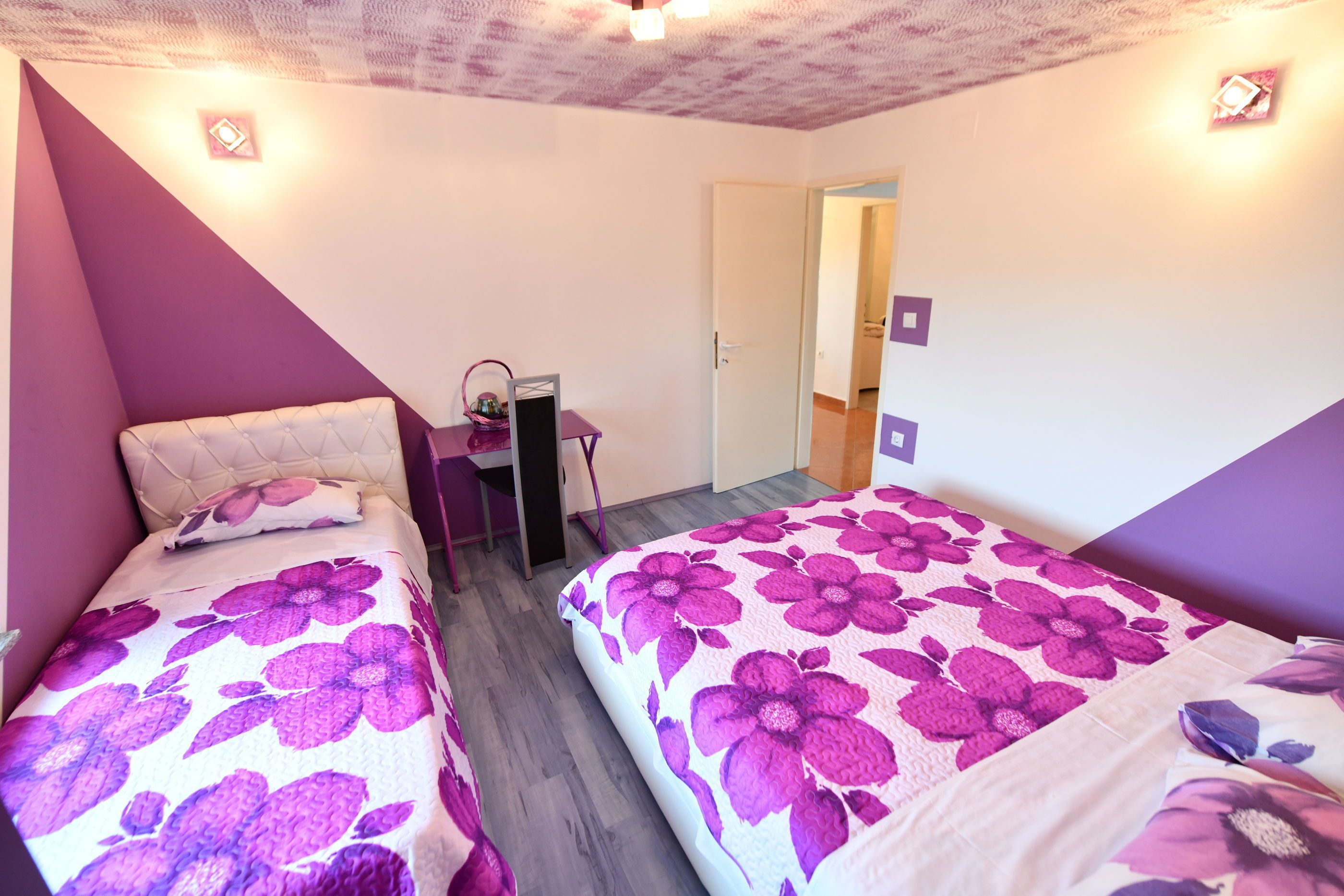 A room with two beds, each with a purple floral bedspread, a desk with a chair, and a door leading to another room.
