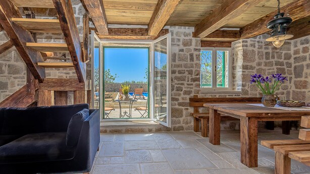 A cozy interior with a stone wall, wooden beams, a black sofa, a wooden table, and a view of the outdoors. - Villa Lipa (Privlaka)