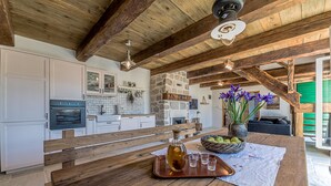 A rustic kitchen with wooden beams, a stone wall, and a wooden dining table with a bottle and glasses. - Villa Lipa (Privlaka)