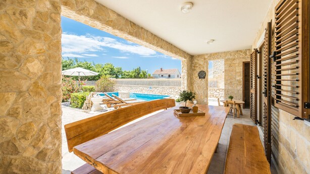 A stone-walled room with a wooden bench and table, a view of a swimming pool, and a door with shutters. - Villa Karin (Privlaka)