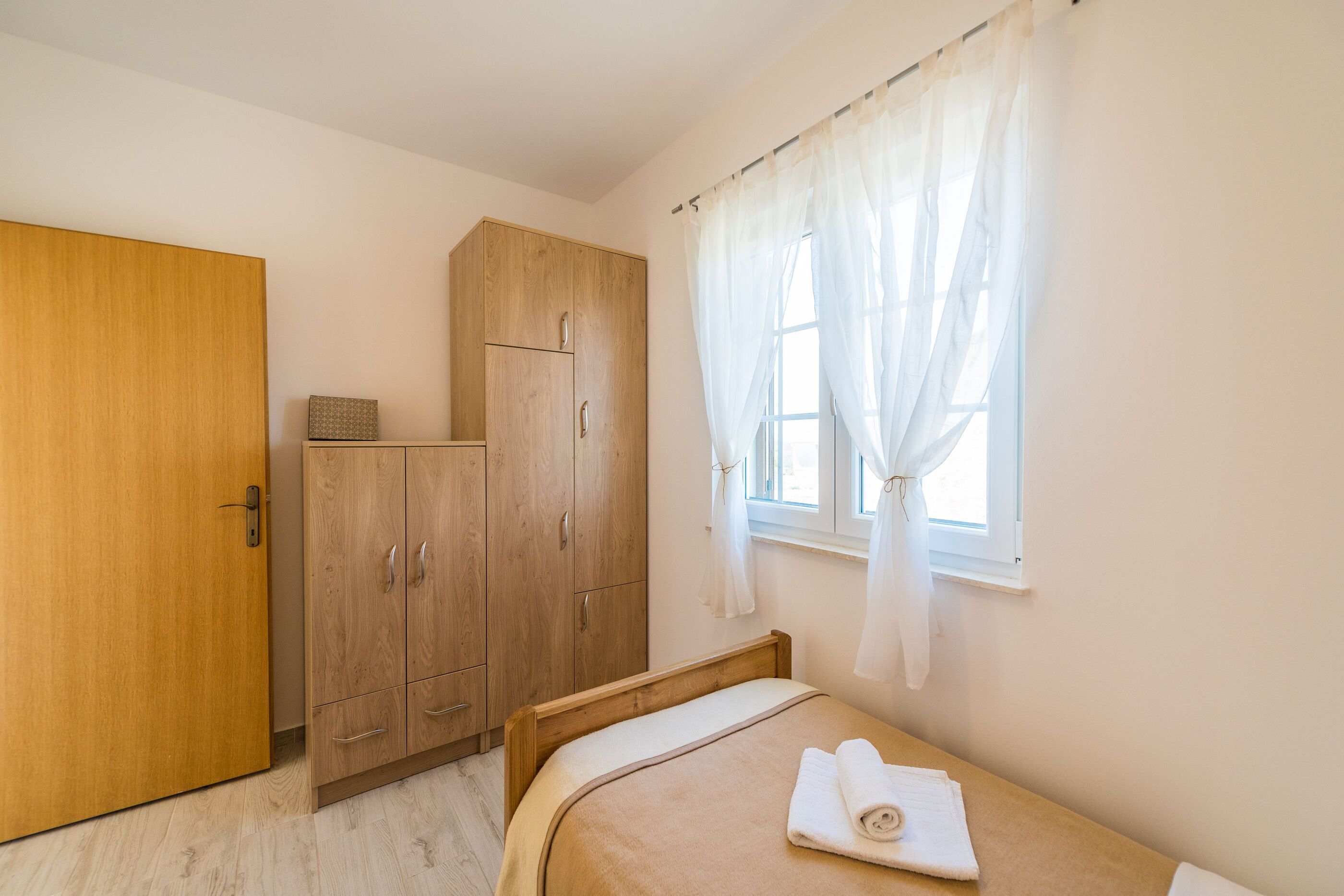 Une chambre avec une armoire en bois, un lit sur lequel est posée une serviette roulée, et une fenêtre ornée de rideaux légers.