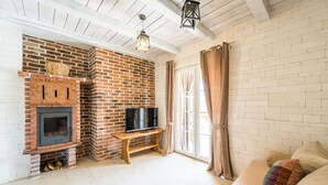 A living room with a brick fireplace, a television on a wooden stand, and a large window with curtains. - Villa Karin (Privlaka)