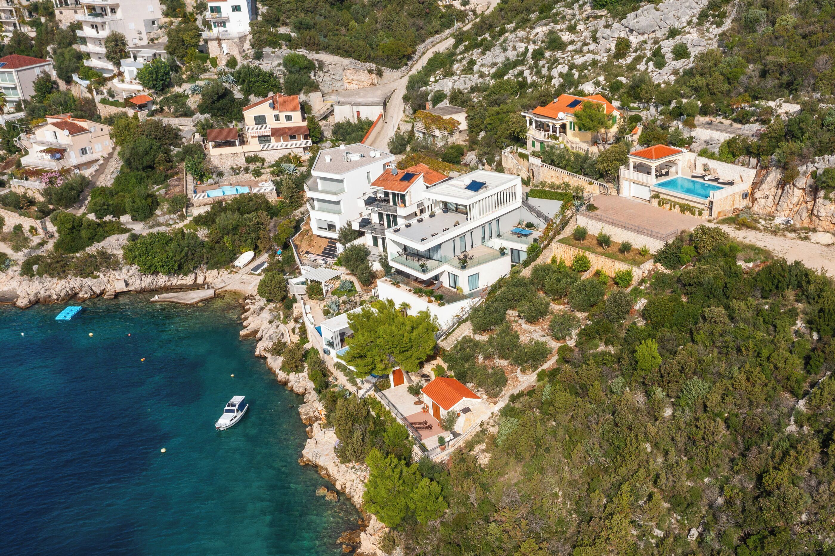 An aerial view of a coastal residential area with modern houses, a swimming pool, and a boat docked near the shore.