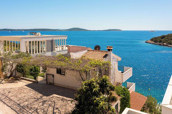 A coastal building with a terrace overlooking a clear blue sea, featuring a mix of modern and traditional architecture.