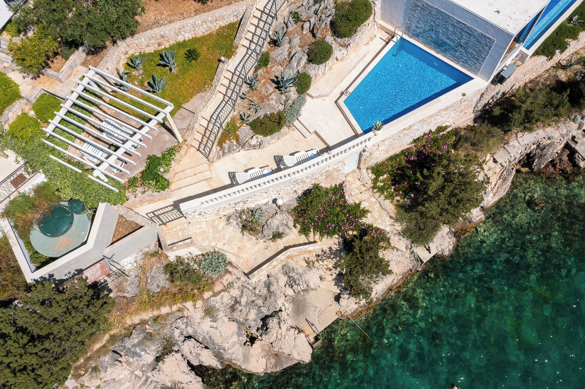 An aerial view of a coastal property with a swimming pool, a staircase leading to a deck, and a rocky shoreline.