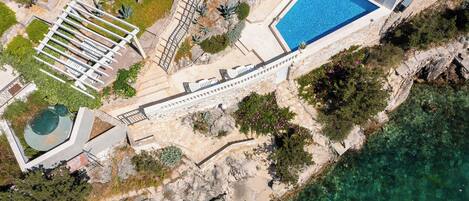 An aerial view of a coastal property with a swimming pool, a staircase leading to a deck, and a rocky shoreline.