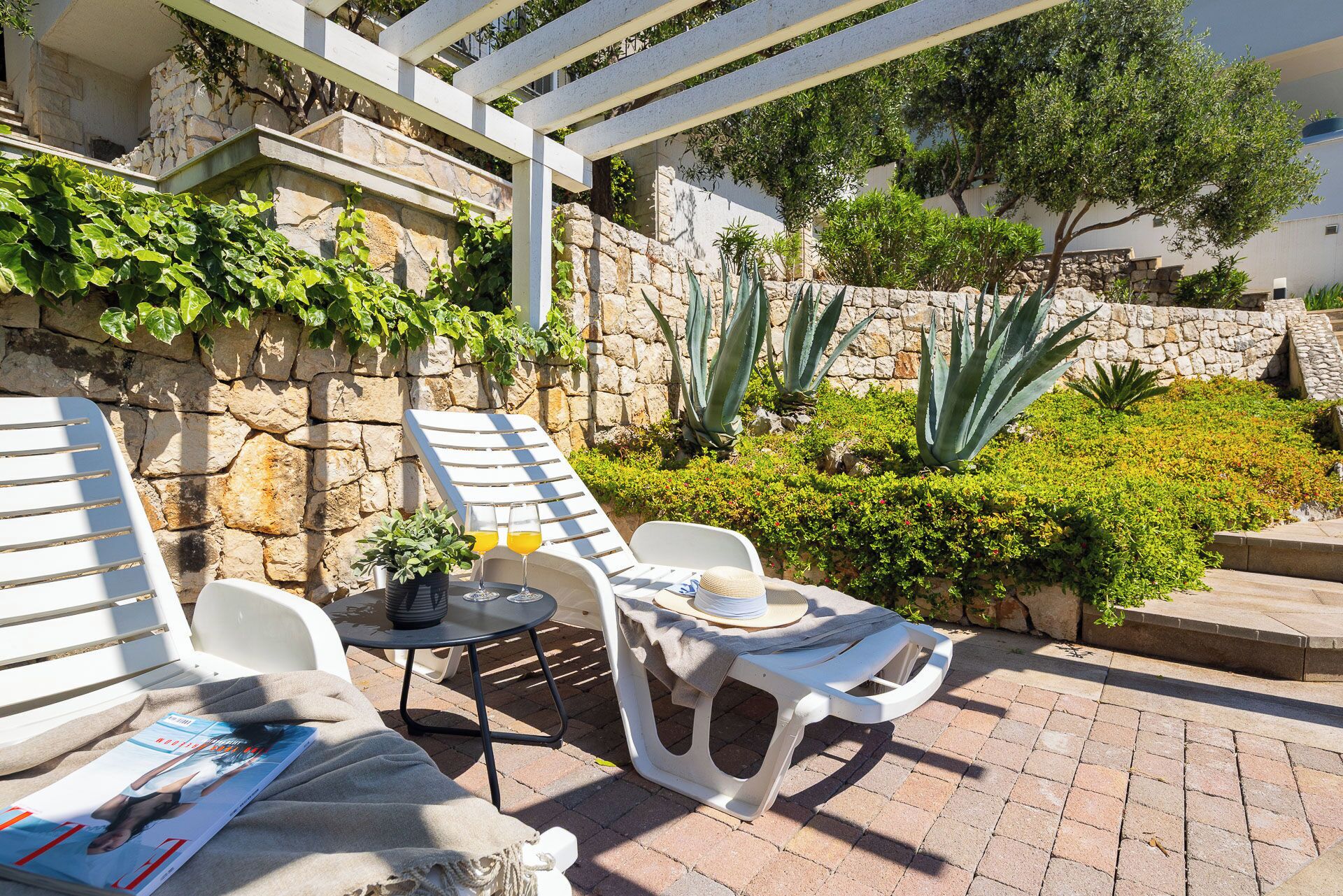 A patio area with white lounge chairs, a small round table, and a magazine. There are plants and a stone wall in the background.