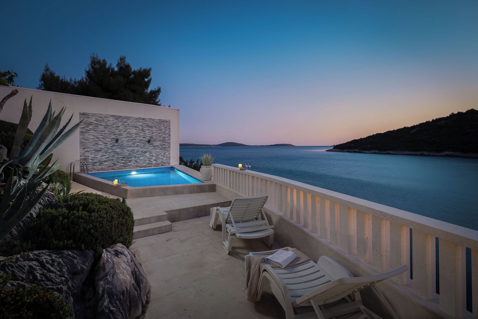 An outdoor swimming pool with lounge chairs and a stone wall, overlooking a body of water and a distant island.