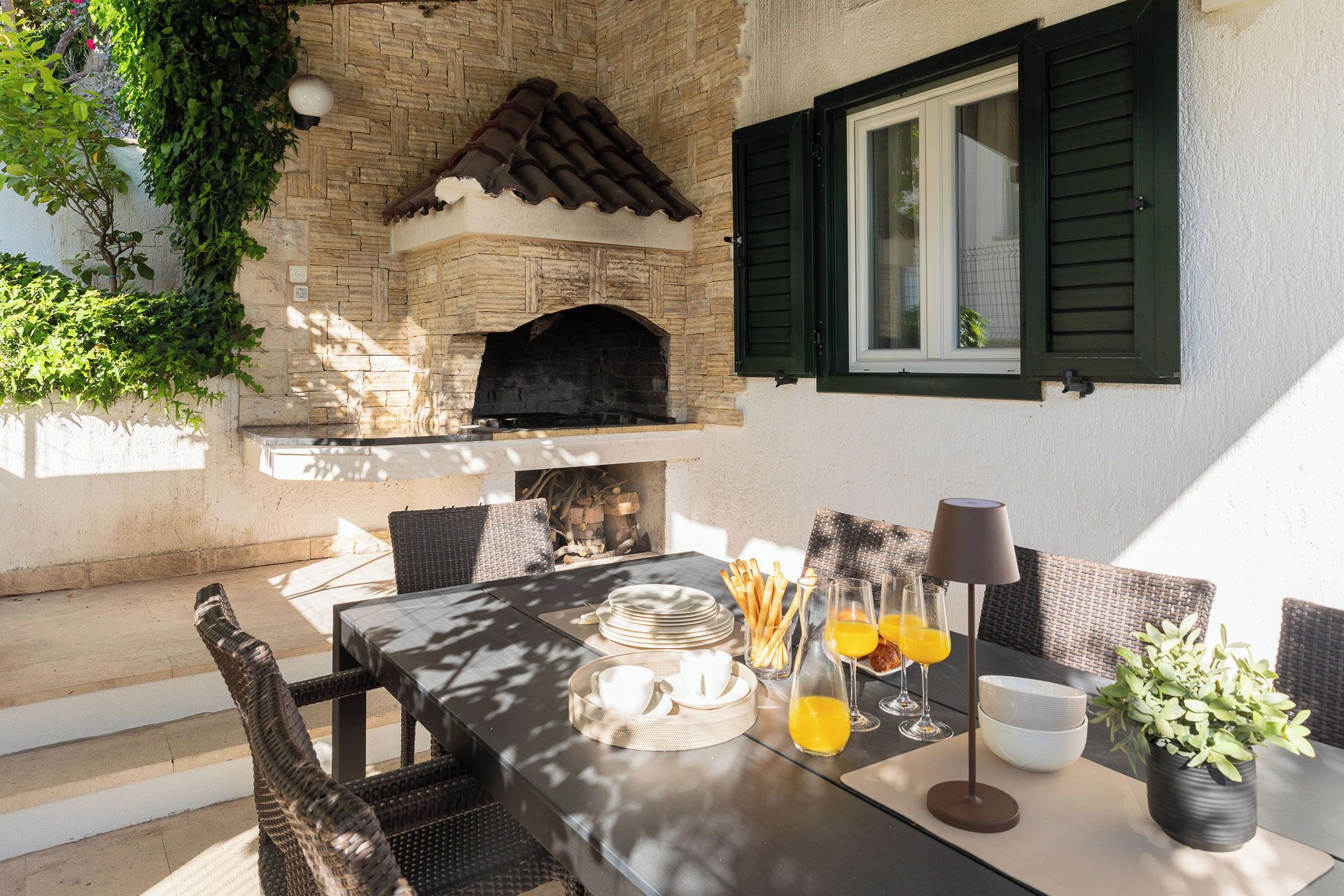 Un espace extérieur aménagé pour les repas, avec une cheminée en pierre, des meubles en osier et une table dressée pour le petit-déjeuner.
