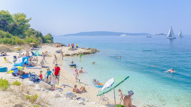 Una spiaggia con acque cristalline, persone che nuotano e vele a distanza.