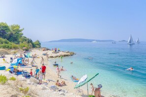 A beach with clear waters, people swimming, and sailboats in the distance.