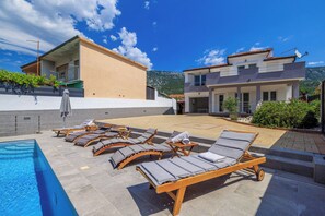 A poolside area with sun loungers and a swimming pool, surrounded by buildings with balconies and a mountainous backdrop.