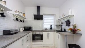 Una cocina moderna con armarios blancos, horno empotrado, microondas y cafetera. Hay ollas colgando y un frutero en la encimera.
