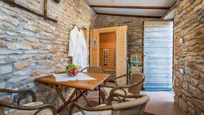 Une pièce aux murs de pierre, avec une table en bois et des chaises en osier, une porte en bois et une petite table sur laquelle sont disposés des fruits.