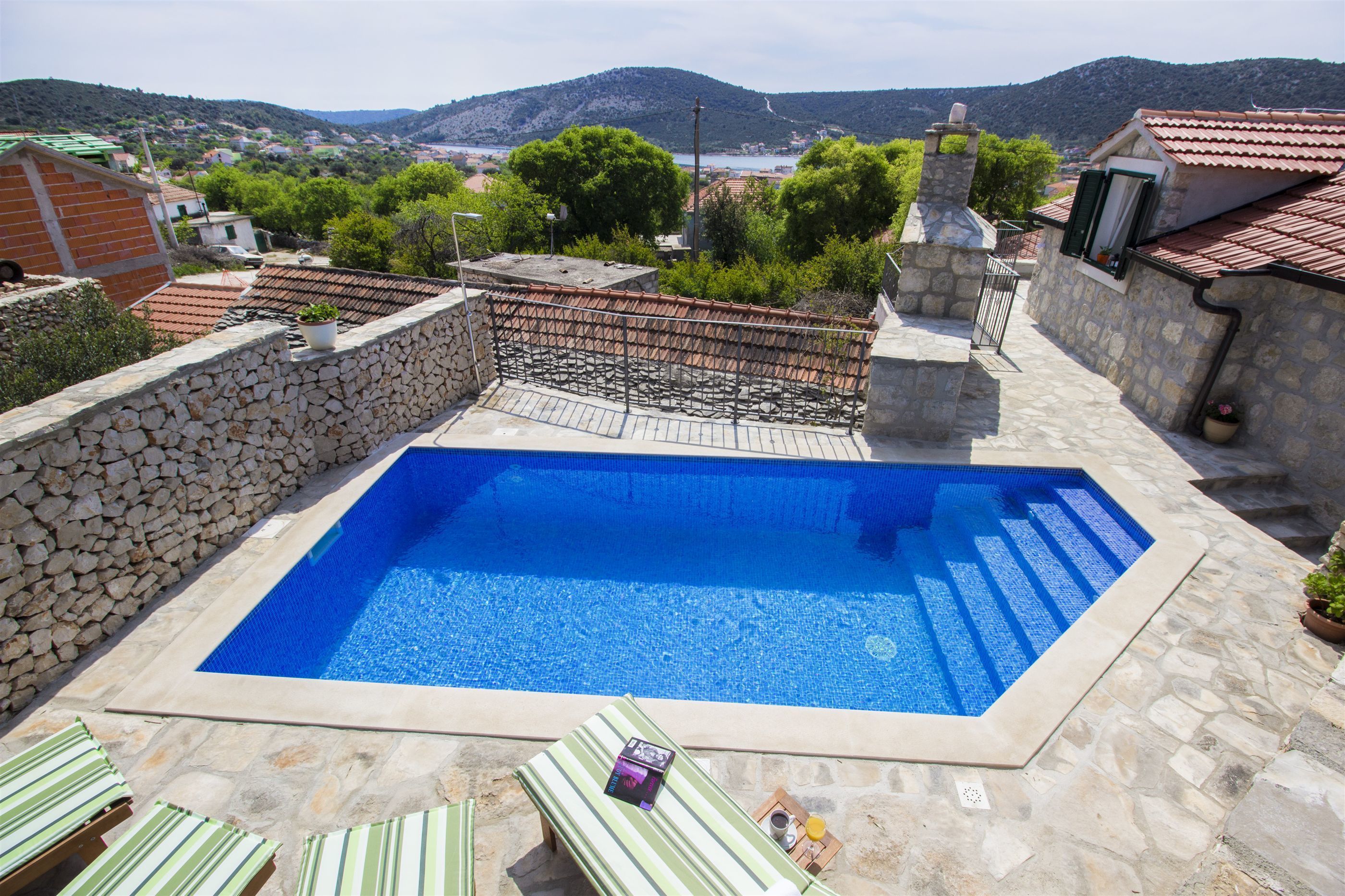 A rectangular swimming pool with a stone border, surrounded by lounge chairs and a stone wall.