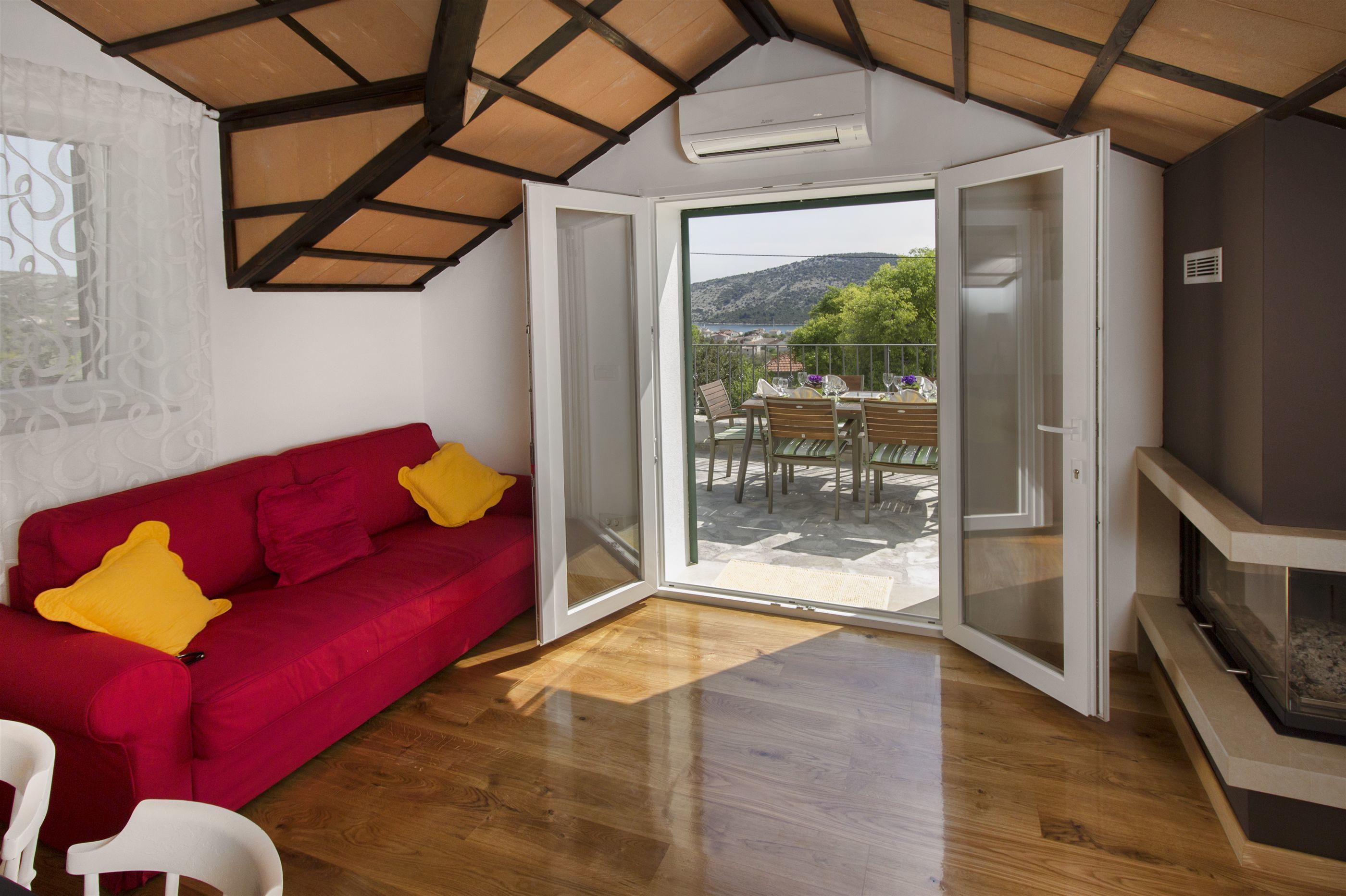 Une pièce avec un canapé rouge, un fauteuil blanc, une cheminée et une porte-fenêtre coulissante donnant sur une terrasse extérieure avec une table et des chaises.