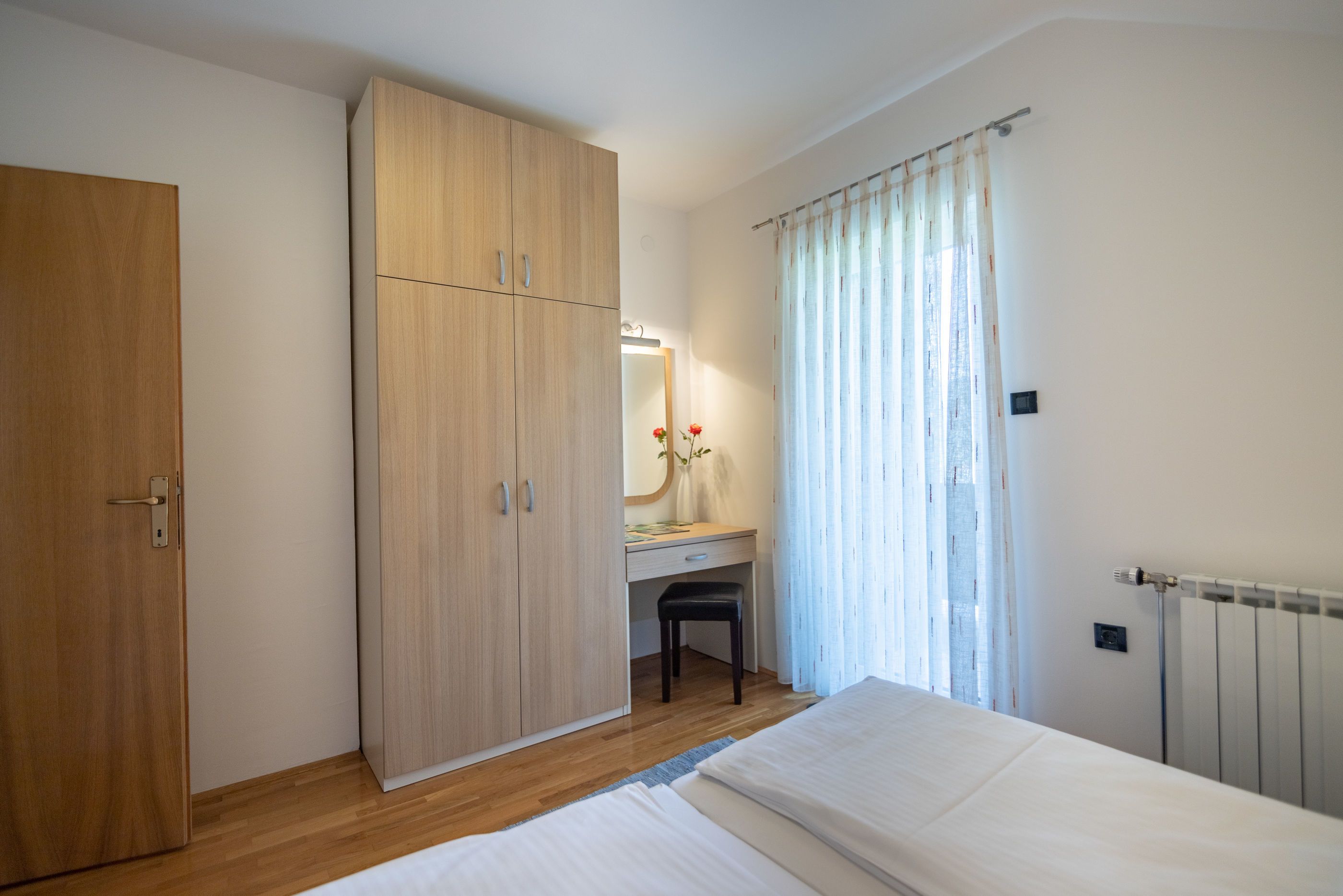 A bedroom with a wooden wardrobe, a desk with a chair, a radiator, and a bed.