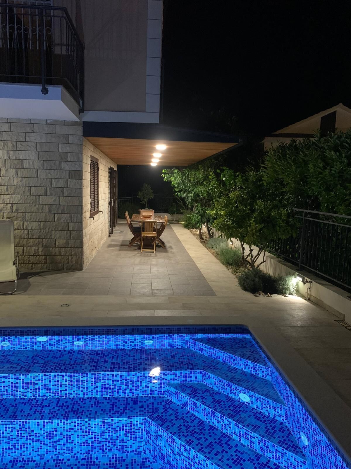 An outdoor swimming pool with a tiled edge, a wooden table and chairs, and a stone wall.