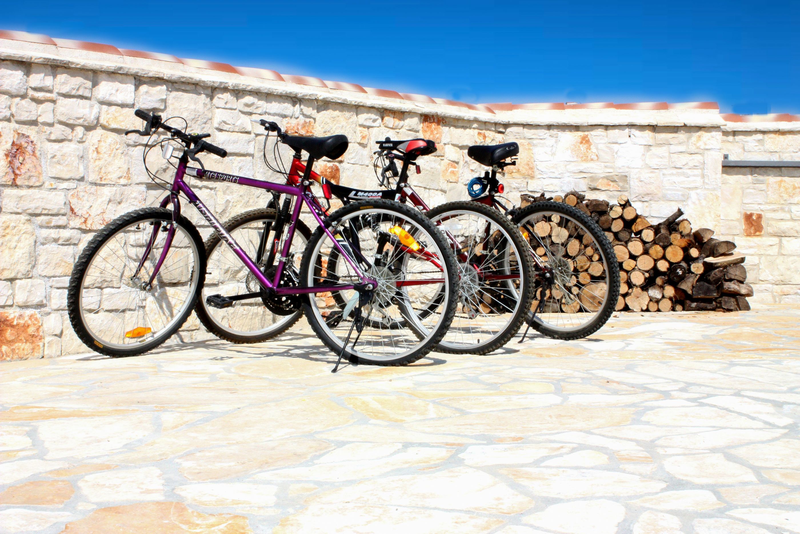 Bicicletas encostadas a uma parede de pedra, com uma pilha de lenha ao fundo.