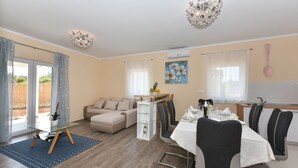 A modern living room with a sofa, dining table, and chairs. There is a glass coffee table, a chandelier, and a painting on the wall. - Villa Rafajela (Privlaka)