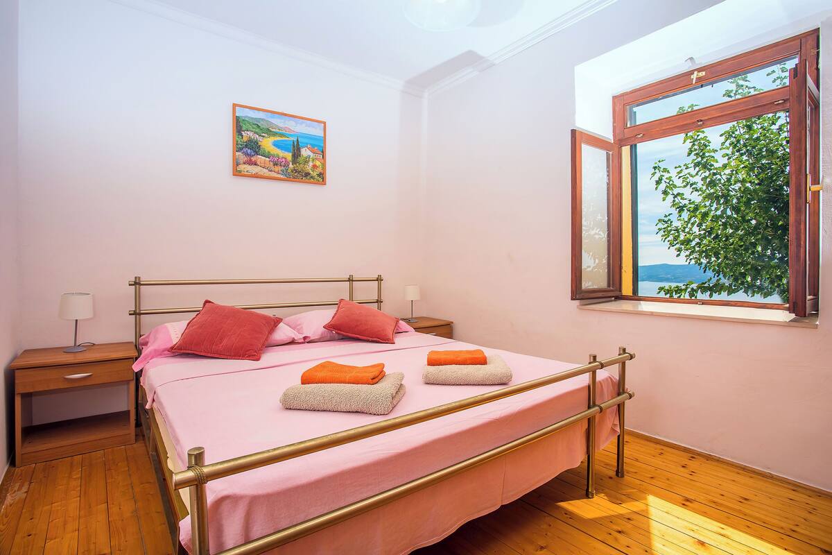 A neatly made bed with pink bedding, a wooden nightstand with a lamp, and a window with a view of greenery.