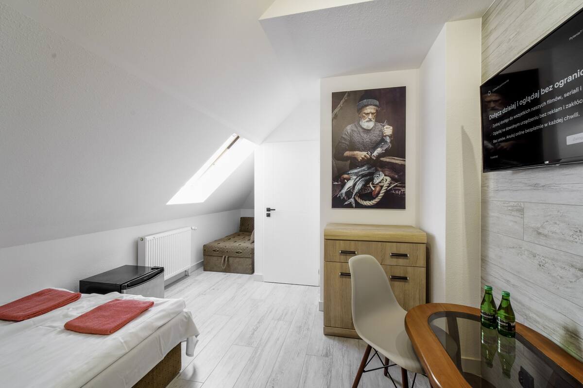 A modern, compact room with a bed, a small table with bottles, a chair, a radiator, a wooden cabinet, and a large poster on the wall.