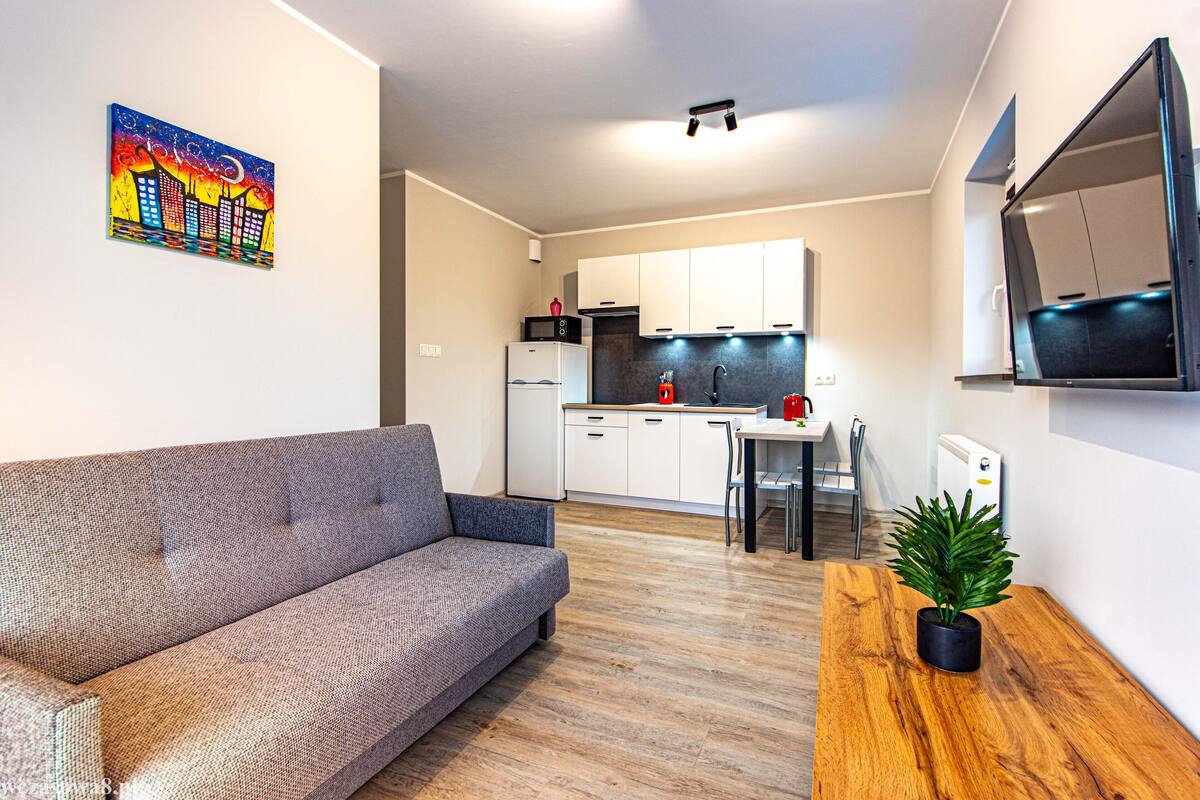 A modern living room with a grey sofa, a wooden dining table with a potted plant, a kitchen with white cabinets, and a colorful cityscape painting on the wall.