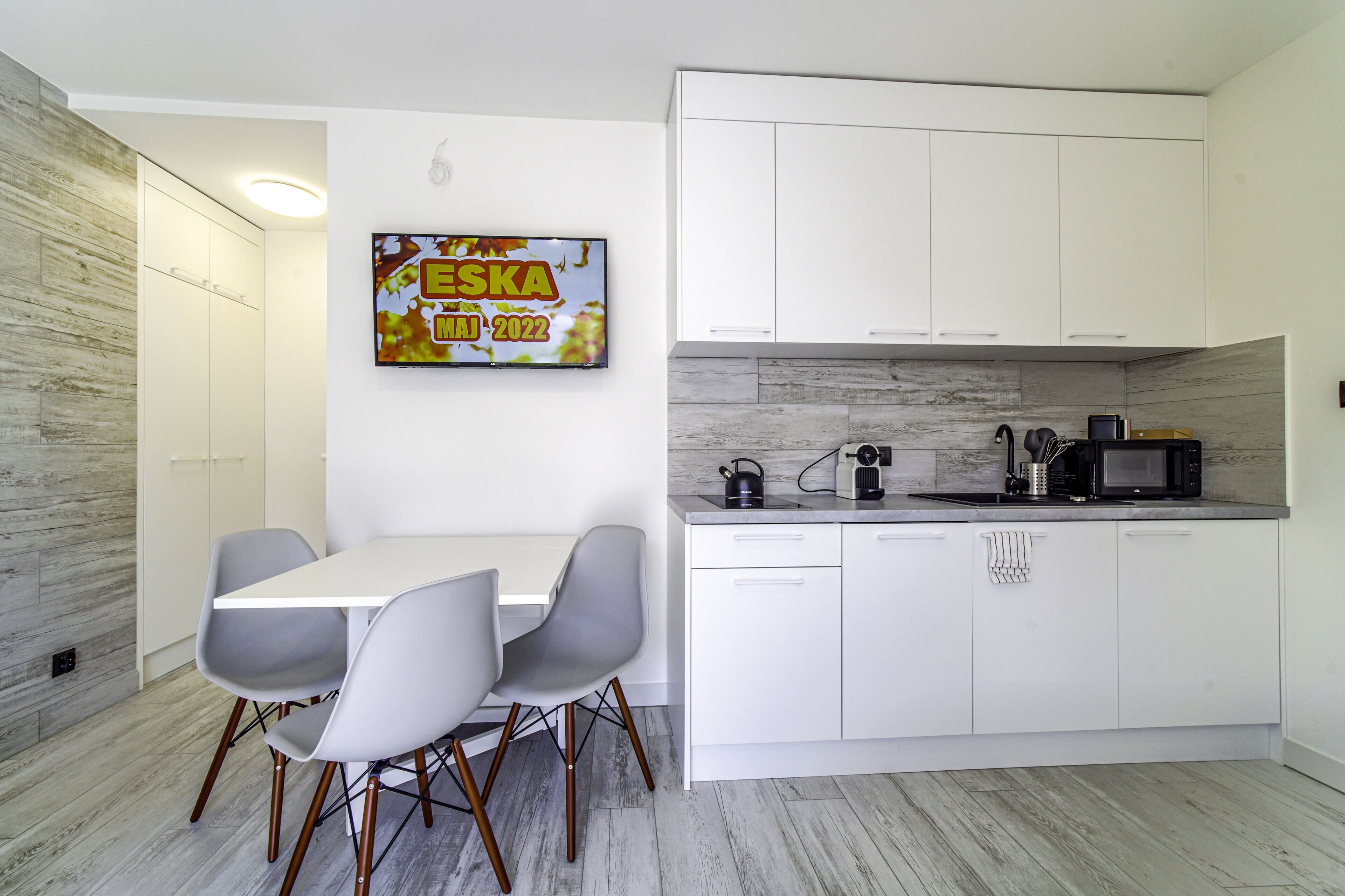 Uma cozinha moderna com armários brancos, uma mesa de jantar com cadeiras e uma TV exibindo 'ESKA MAI 2022'.