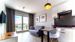 A modern living room with a sofa, a dining table, and chairs. There is a TV mounted on the wall, a kitchen area with cabinets and a stove, and a large window with curtains. - Wczasowa 8 Apartments Sarbinowo (Sarbinowo)