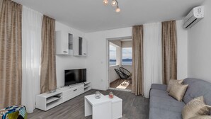 En moderne stue med en grå sofa, et hvidt tv-møbel og et fjernsyn. Der er et vindue med gardiner, en dør til udendørs og en loftlampe.