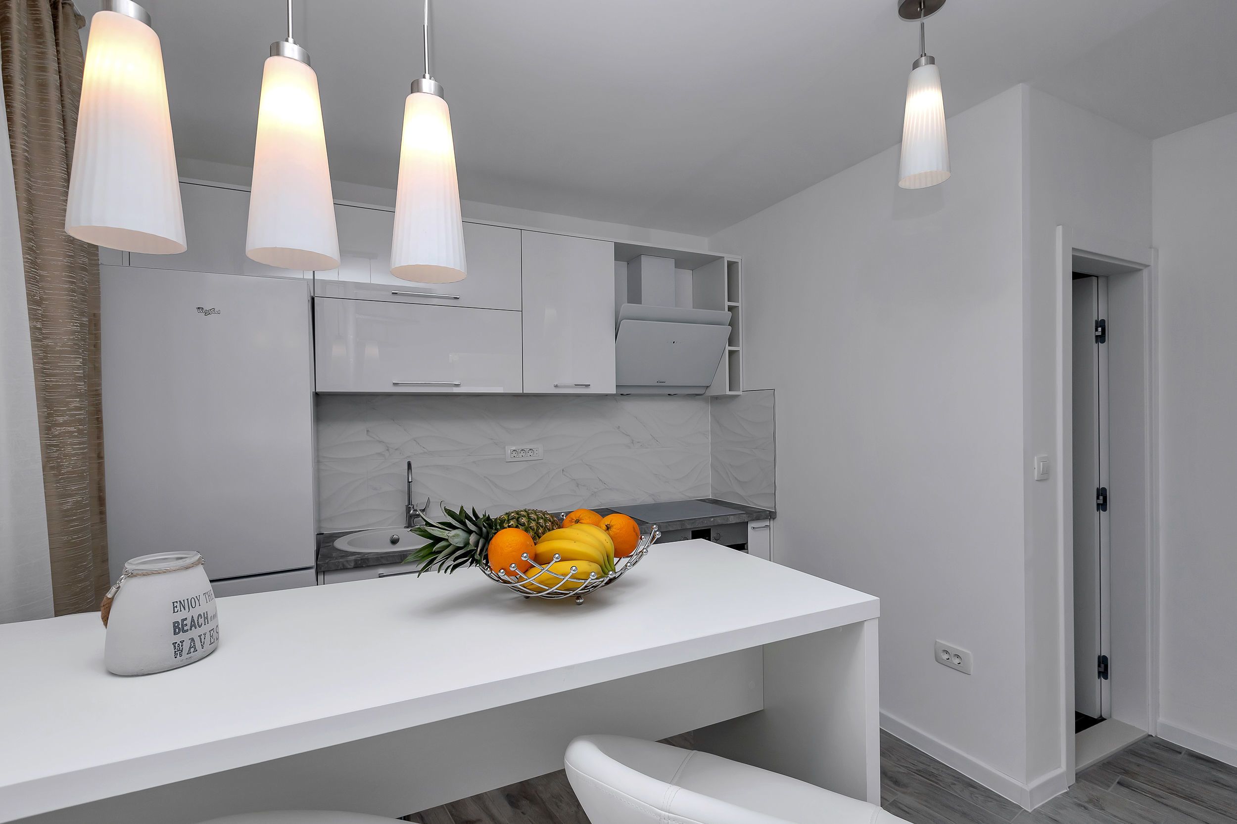 Une cuisine moderne avec des meubles blancs, un panier de fruits sur le comptoir et des luminaires suspendus.
