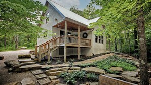 A rustic cabin with a wooden deck and a porch, surrounded by a forested landscape.