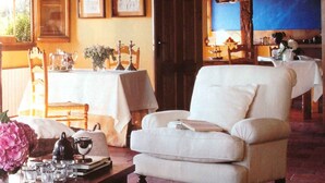 A cozy room with a wooden ceiling, a white armchair, a table with a vase of flowers, and a view into another room with a table set for dining. - Casa Rota (Arahuetes)