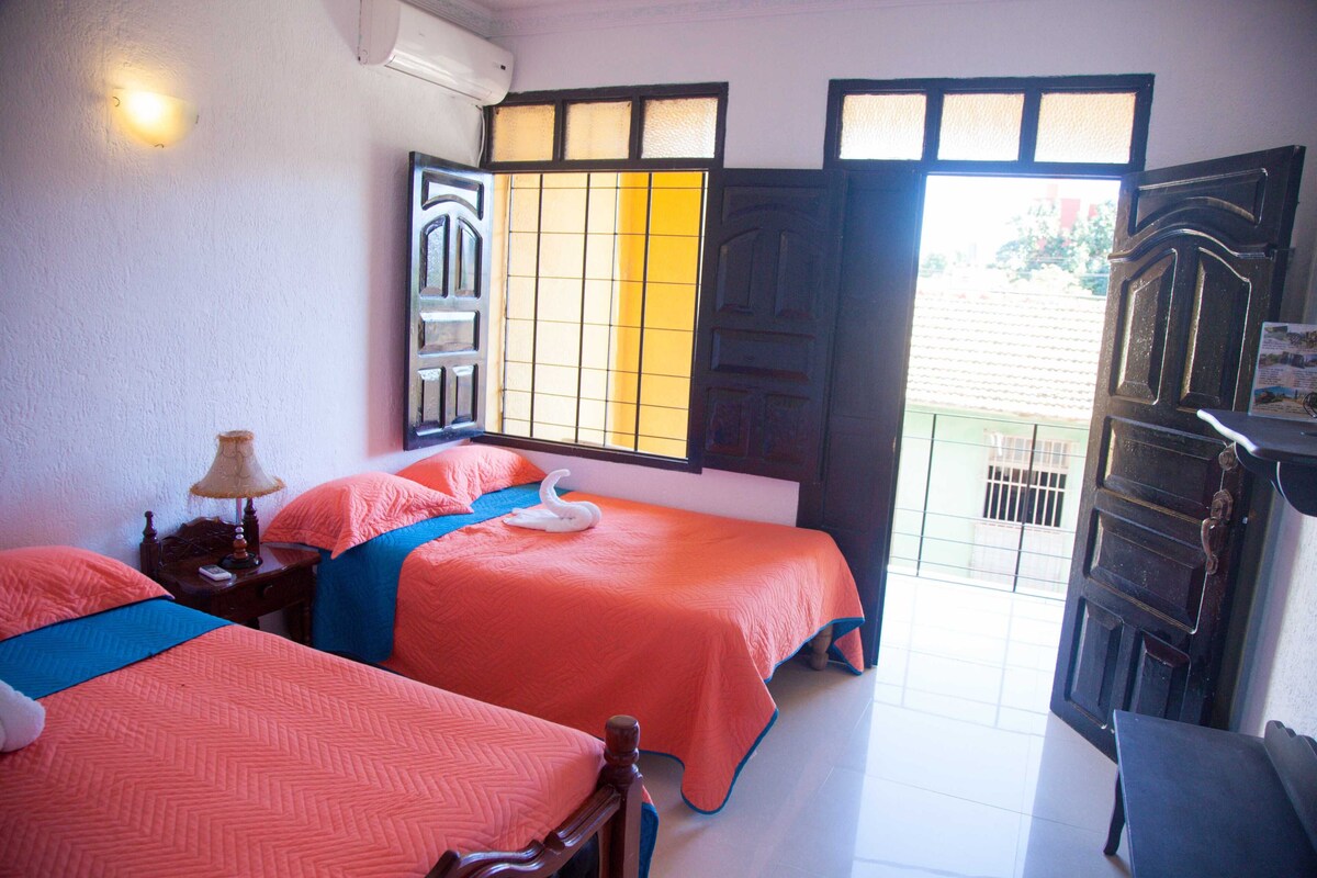 A room with two beds, a window with a view, a door, and a small table with a lamp.