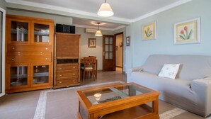 A living room with a glass-top coffee table, a sofa, wooden cabinets, and a dining area with a table and chairs. - Apartamento Paseo Maritimo 27 (Cádiz)
