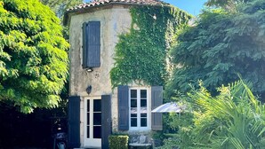 A rustic stone building with a tiled roof, a small porch, and a garden area with a bench and umbrella. - Gîte La Folie de Labahou (Anduze)